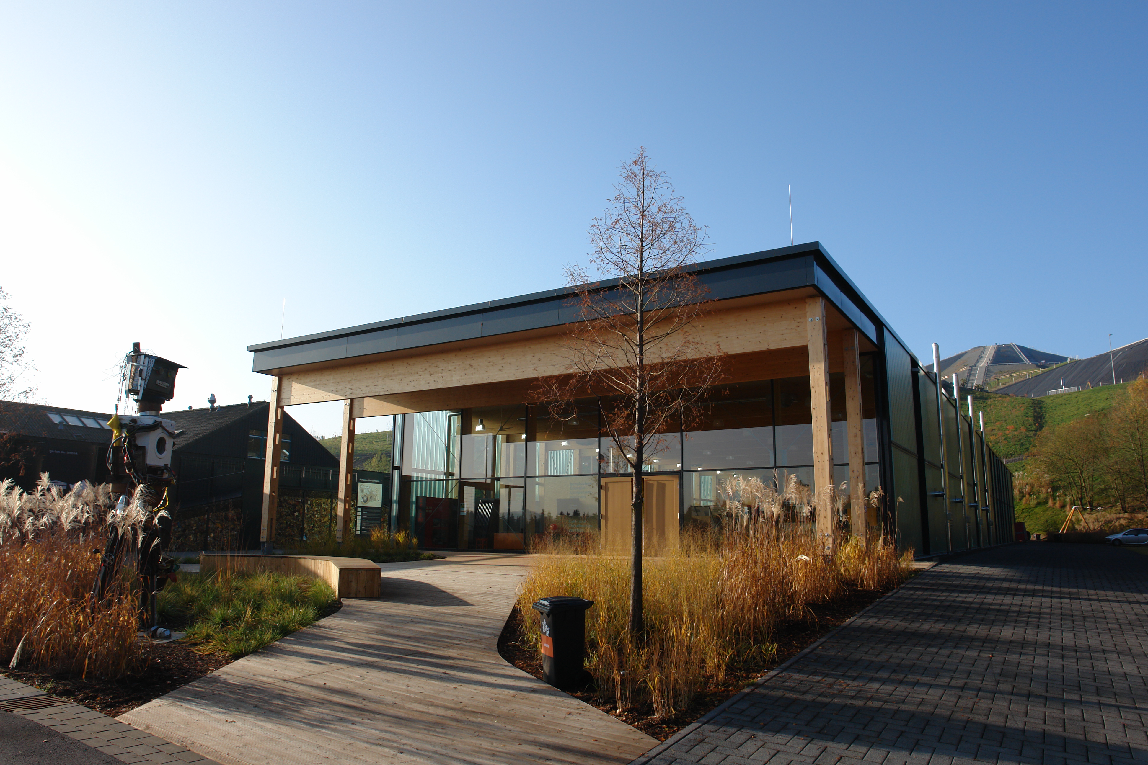 Modernes Gebäude mit Holzelementen und Glasfront, umgeben von Gräsern und einem Baum. Links eine Skulptur, im Hintergrund ein grüner Hügel.