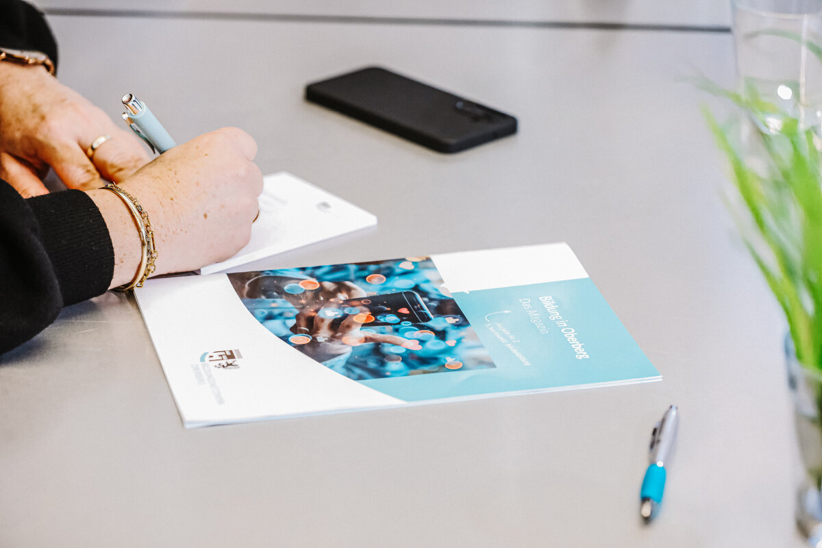 Eine Person schreibt mit einem Stift auf Papier. Auf dem Tisch liegen ein Magazin mit einem blauen Titelblatt, ein schwarzes Smartphone und ein weiterer Stift.
