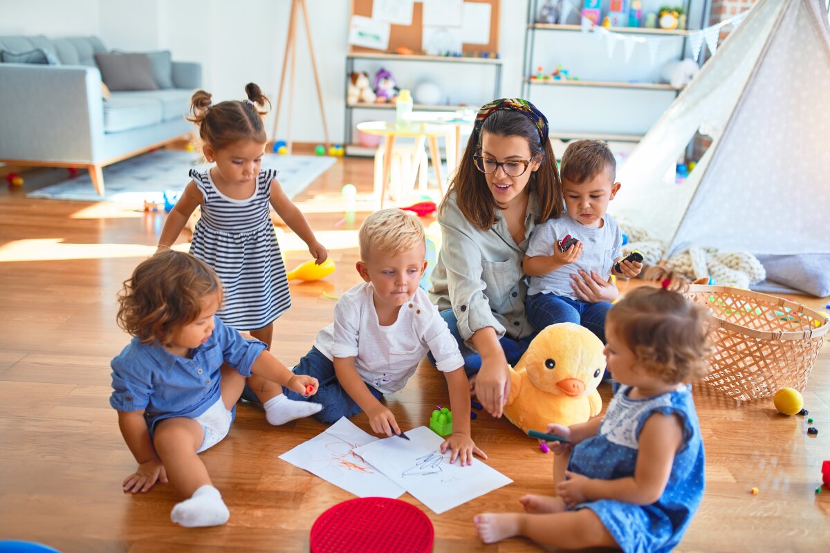 Beautiful teacher and group of toddlers sitting on the floor drawing using paper and pencil around lots of toys at kindergarten