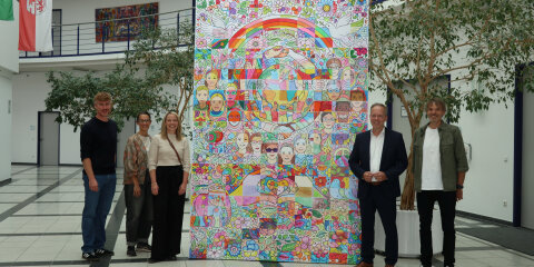 Fünf Personen stehen in einer modernen Halle vor einem großen, bunten Kunstwerk mit Motiven wie Regenbögen, Blumen, Tauben und Gesichtern. Im Hintergrund sind Pflanzen, Flaggen und Türen zu sehen.