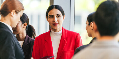Professionals converse at a networking event; a woman in a red blazer speaks with colleagues near large windows while another uses a tablet.