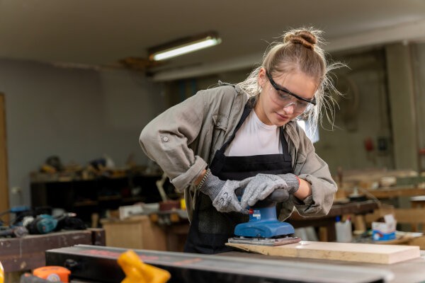 Eine Person in Arbeitskleidung und Schutzbrille schleift mit einem elektrischen Schleifgerät ein Stück Holz in einer Werkstatt. Im Hintergrund sind Werkzeuge und Arbeitsflächen sichtbar.