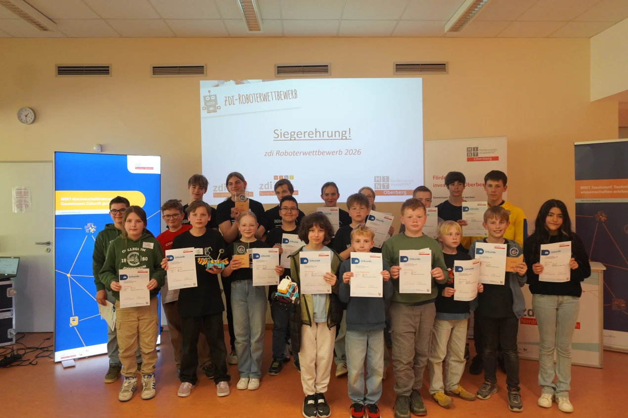 Eine Gruppe von Kindern und Jugendlichen hält Urkunden und steht vor einer Leinwand, auf der 'zdi Roboterwettbewerb 2026 - Siegerehrung!' angezeigt wird.