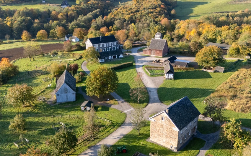 Luftaufnahme eines historischen Dorfes mit mehreren Fachwerk- und Steinhäusern, einer Scheune und grünen Wiesen. Umgeben von herbstlichen Bäumen.