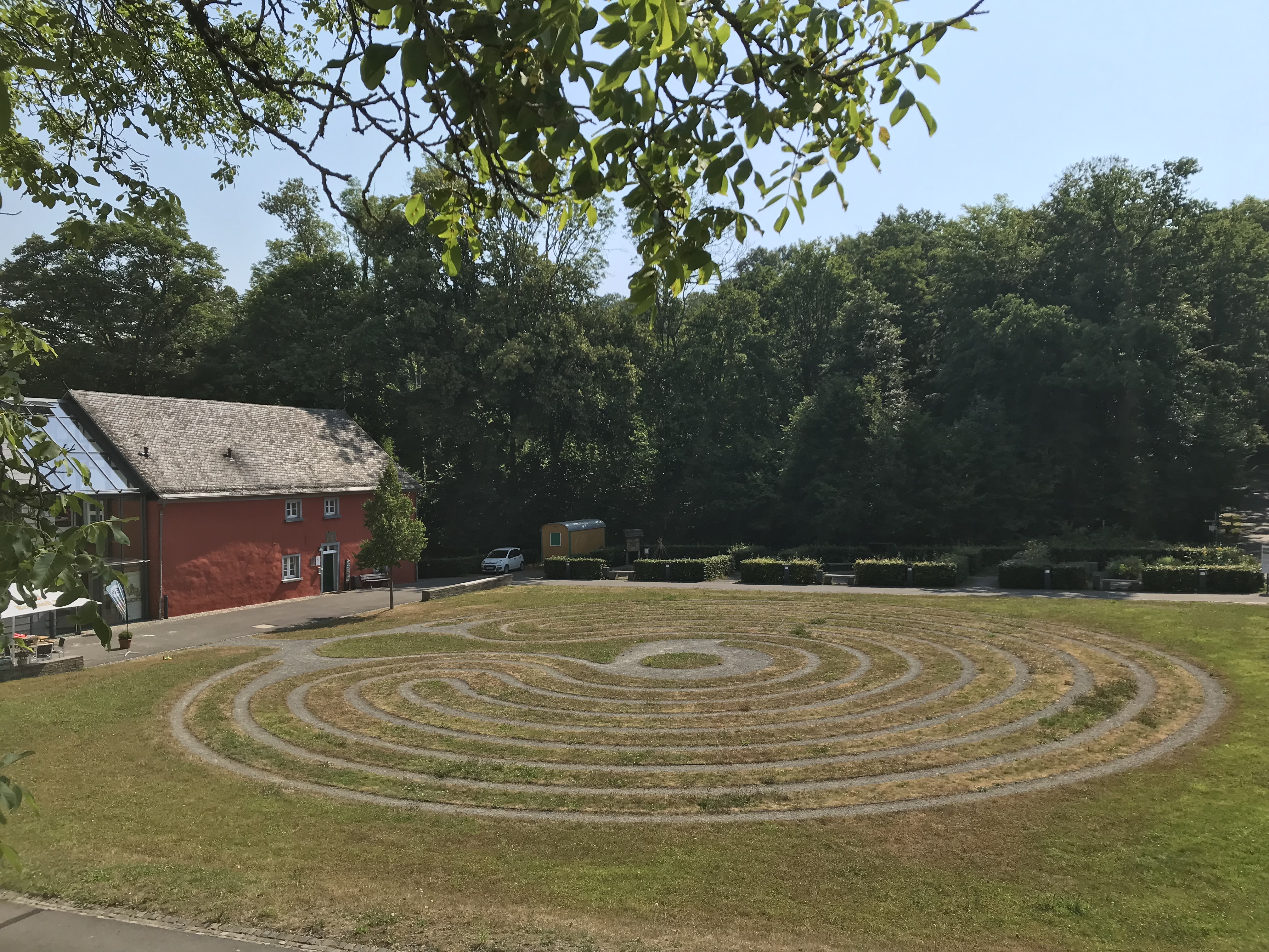 Ein rotes Gebäude mit einem grauen Dach steht links im Bild. Davor befindet sich eine kreisförmige Rasenfläche mit einem labyrinthartigen Muster. Im Hintergrund sind Bäume und ein blauer Himmel zu sehen.