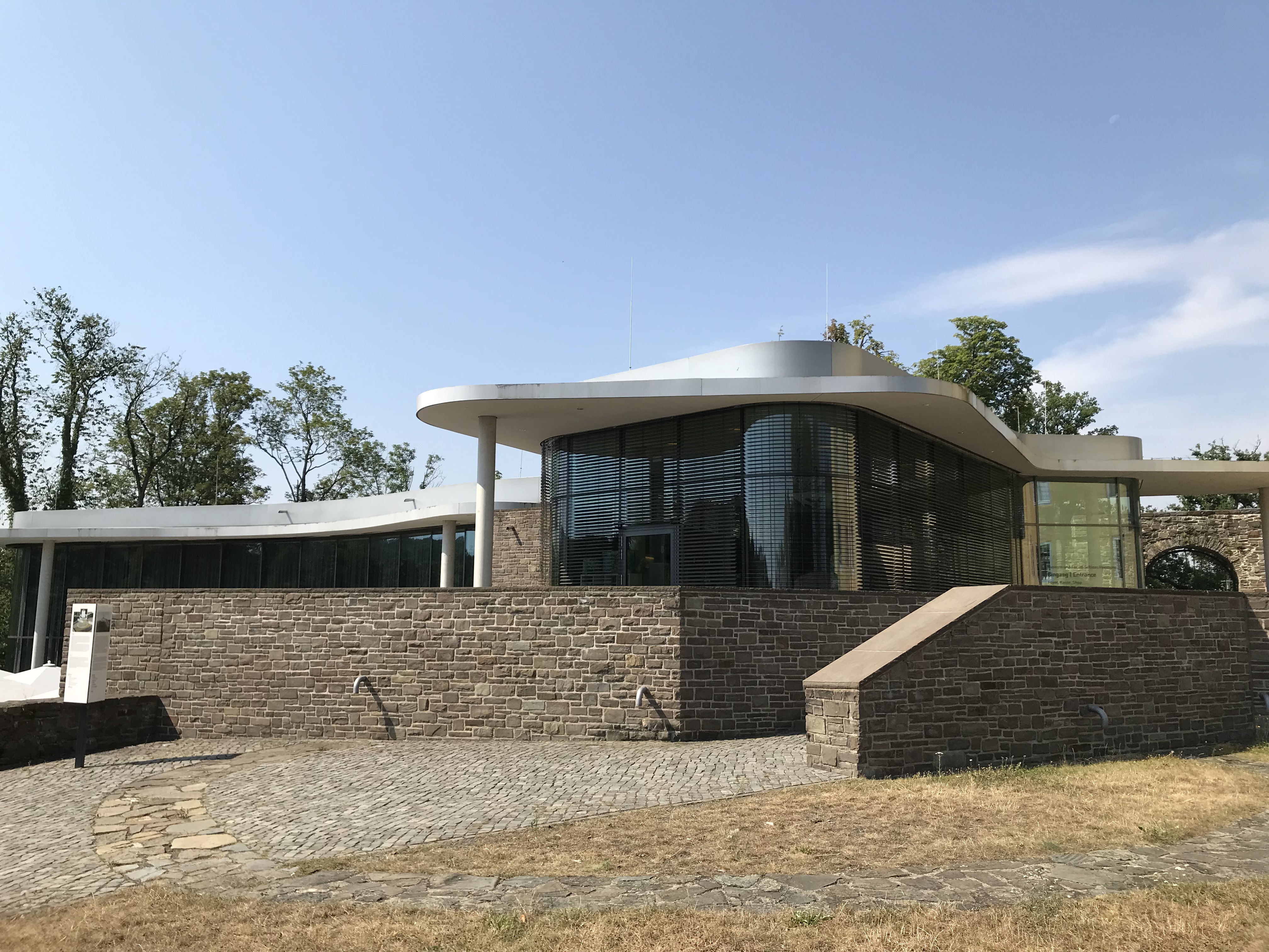 Modernes Gebäude mit geschwungenem, weißem Dach, großen Glasfronten und einer Fassade aus Naturstein. Davor gepflasterter Weg und Rasenfläche.