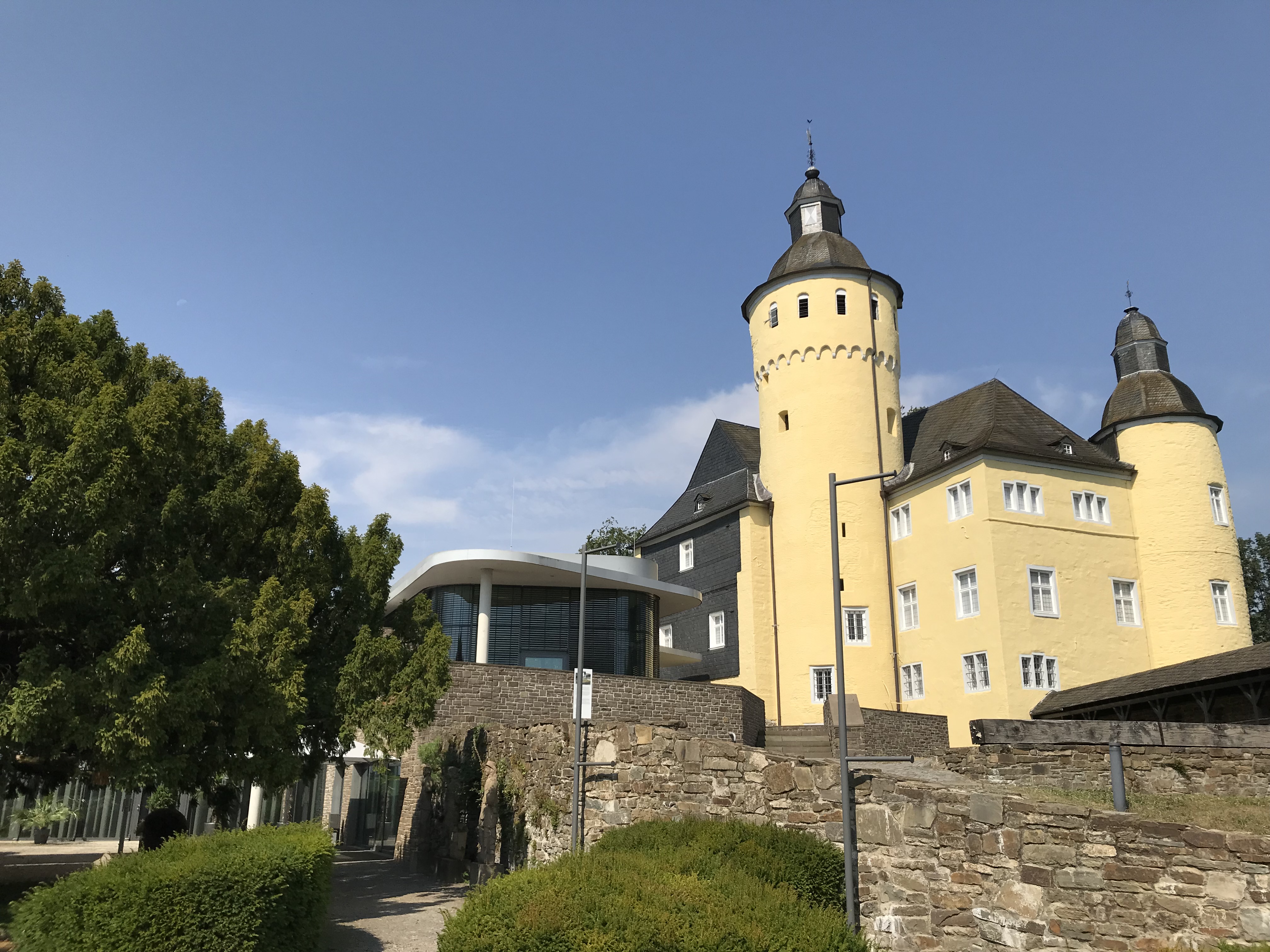 Historisches gelbes Gebäude mit runden Türmen und spitzen Dächern, flankiert von einem modernen Gebäude mit Glasfassade und einem Baum im Vordergrund. Klare, blaue Himmel im Hintergrund.