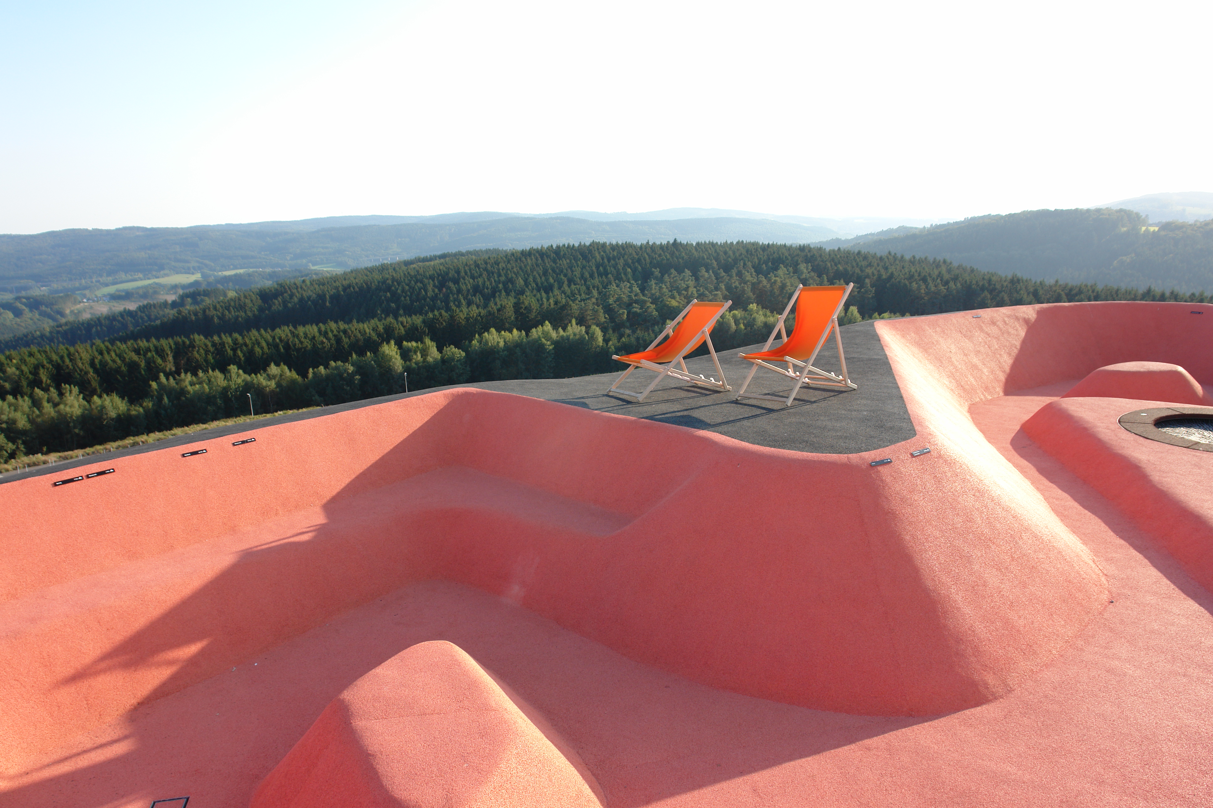 Zwei orange Liegestühle stehen auf einer schwarzen Fläche mit Blick auf eine hügelige Landschaft und einen Wald im Hintergrund. Im Vordergrund befinden sich geschwungene, rosa gefärbte Strukturen.