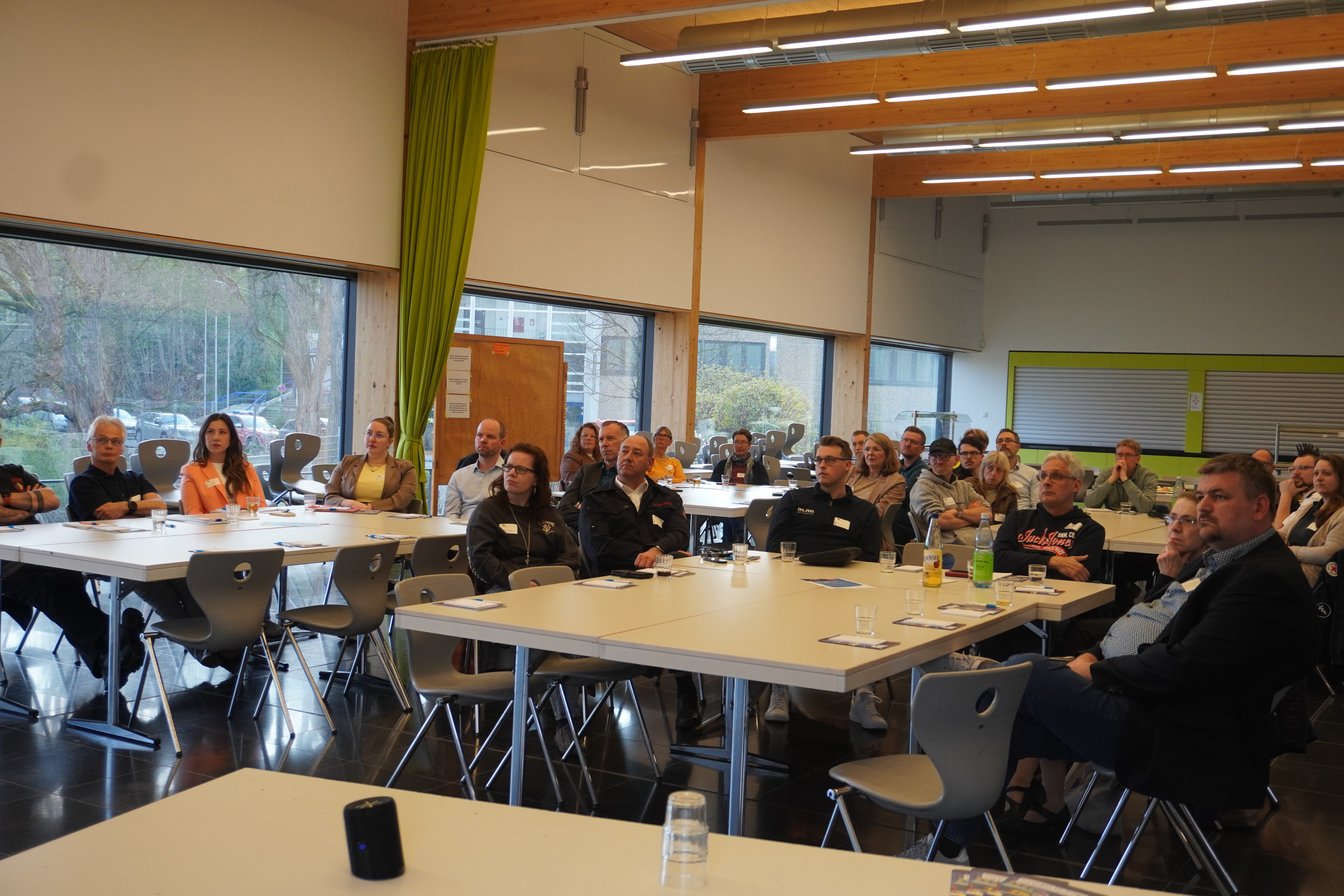 Ein Gruppenfoto von Menschen, die an Tischen in einem hellen Raum sitzen. Im Hintergrund sind große Fenster und grüne Vorhänge sichtbar.
