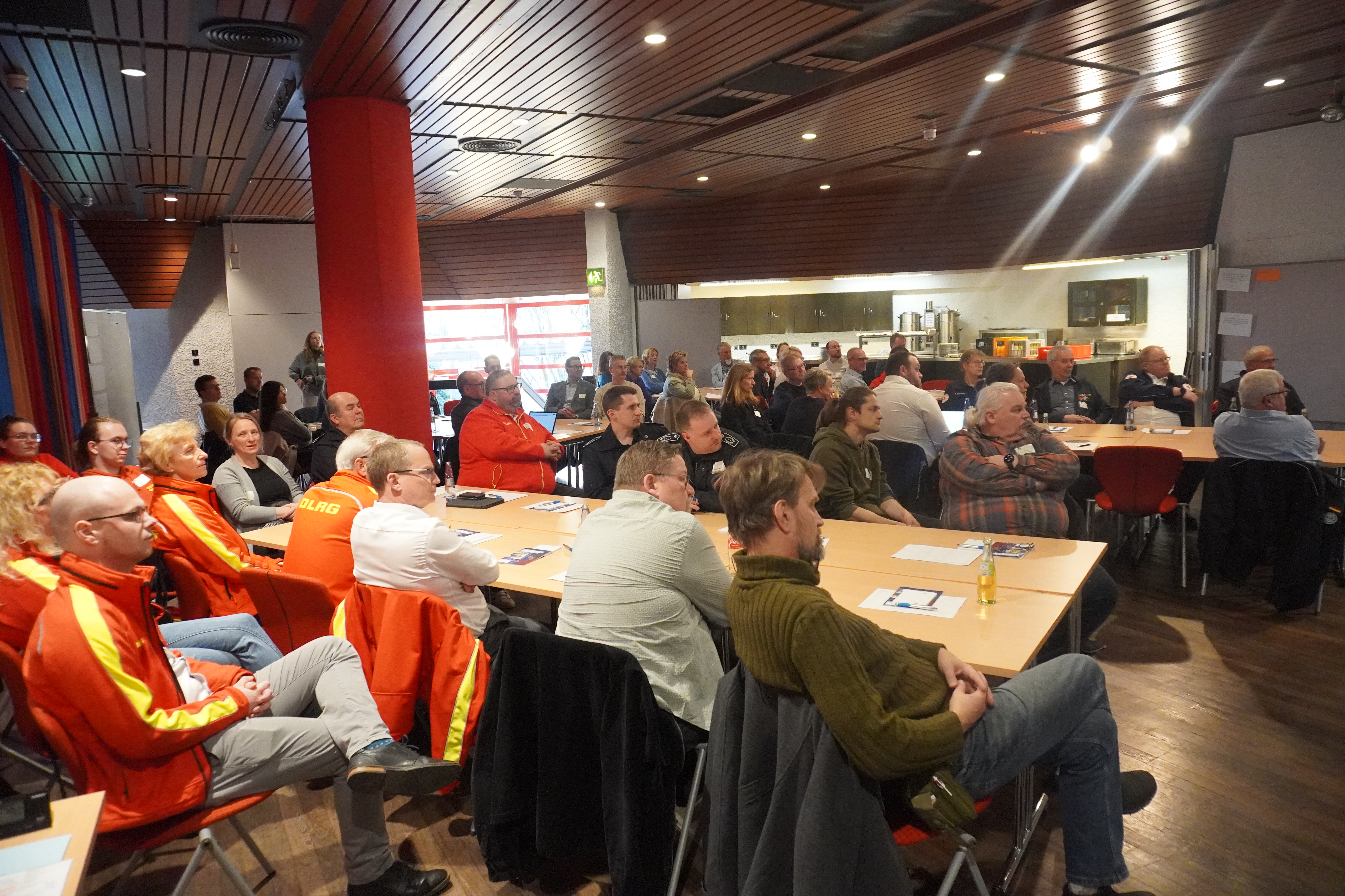 Eine Gruppe von Menschen sitzt in einem Raum an Tischen und hört aufmerksam zu. Einige tragen orangefarbene Jacken. Im Hintergrund sind eine Küche und bunte Vorhänge zu sehen.