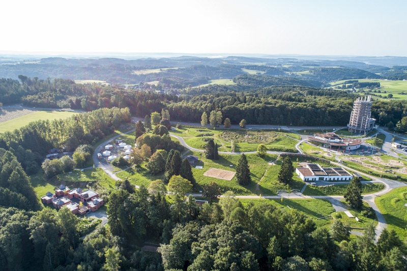 Luftaufnahme einer grünen Landschaft mit Bäumen, Straßen und Gebäuden. Rechts ist ein hoher runder Turm und weitere Gebäude sichtbar.