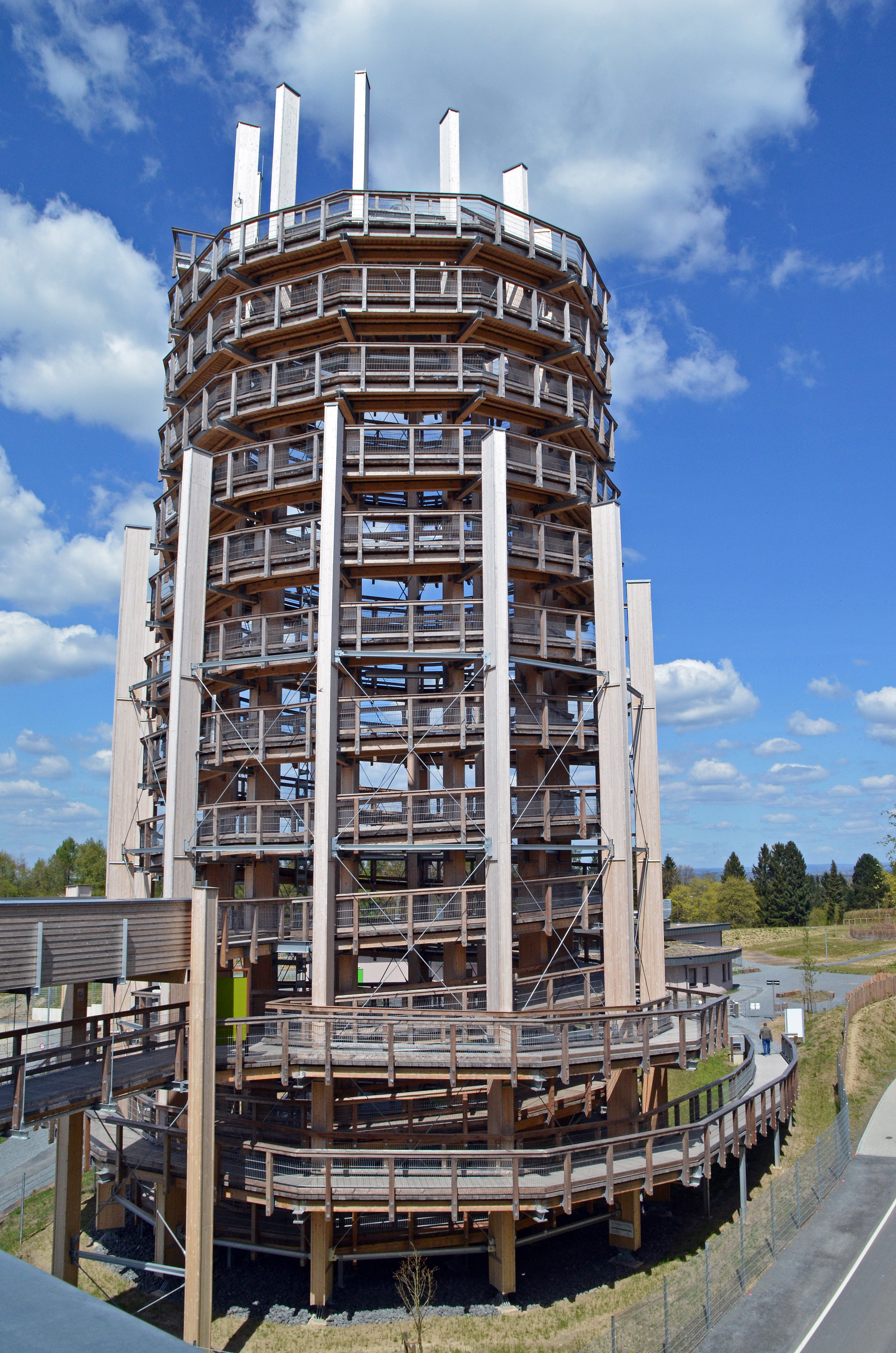 Ein hoher, runder Aussichtsturm aus Holz mit mehreren Plattformen und Geländern. Der Turm hat eine spiralförmige Rampe und steht in einer offenen Landschaft unter blauem Himmel mit Wolken.