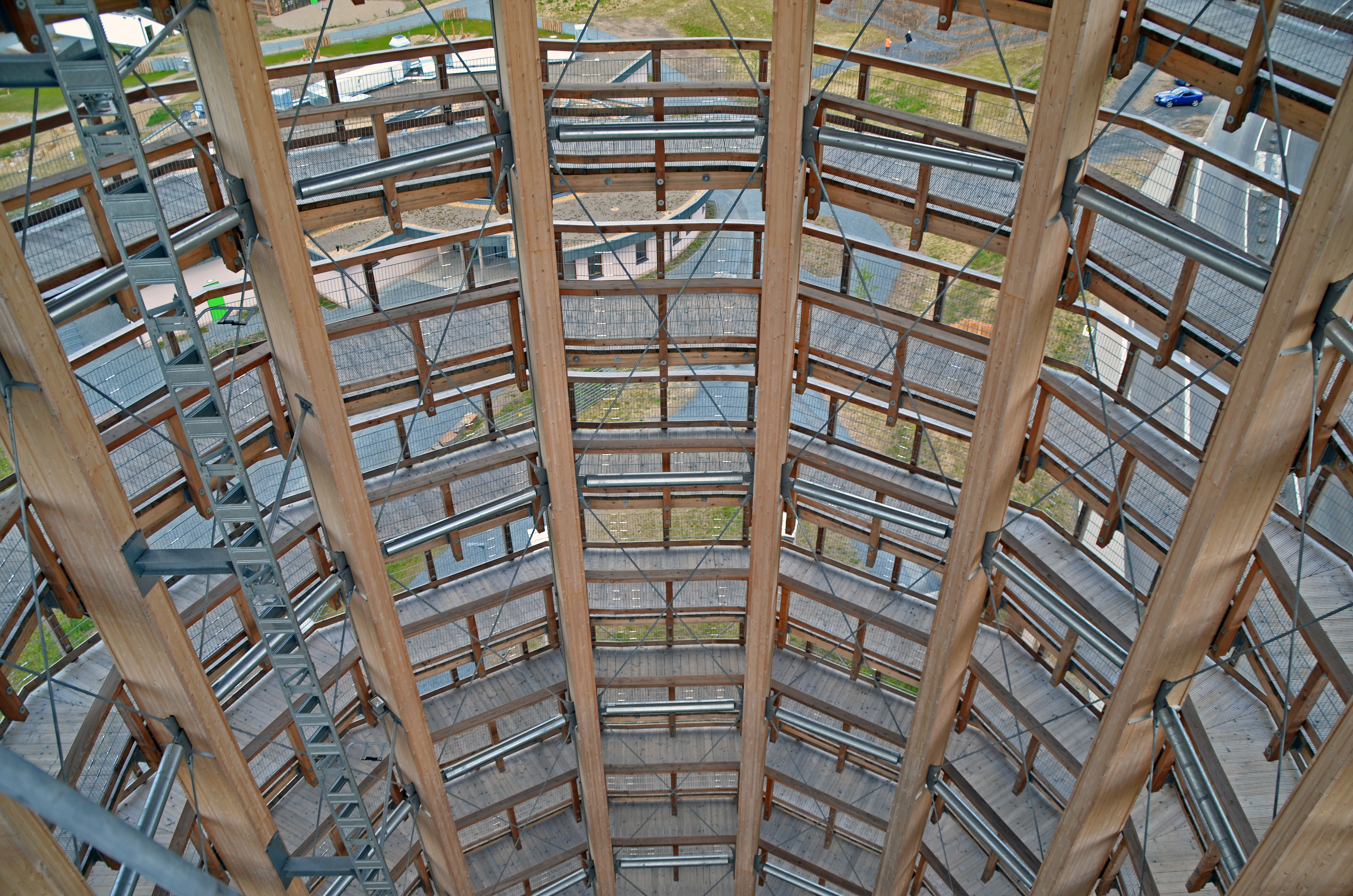 Blick von oben auf die spiralförmige Konstruktion eines Holz-Aussichtsturms mit Stufen und Geländern aus Holz und Metall. Im Hintergrund ist eine Landschaft mit Straßen und Rasenflächen zu sehen.