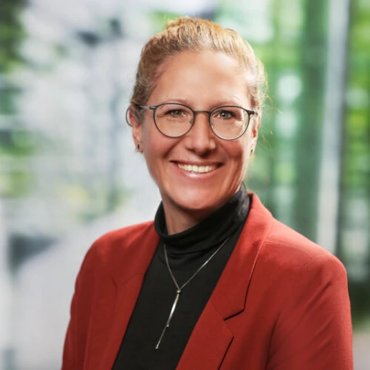 Eine lächelnde Person mit Brille und roten Haaren, die zu einem Knoten gebunden sind, trägt ein rotes Sakko und einen schwarzen Rollkragenpullover. Der Hintergrund ist unscharf mit grünen und weißen Tönen.