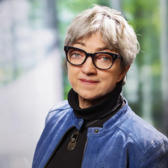 Eine Frau mit grauem, kurzen Haar trägt eine schwarze Brille, eine blaue Jacke und einen schwarzen Rollkragenpullover. Der Hintergrund ist unscharf und zeigt Grünflächen.