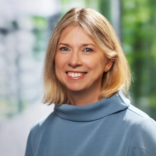 Porträt einer lächelnden Person mit schulterlangem blondem Haar in einem blauen Oberteil vor einem unscharfen, grünen Hintergrund.