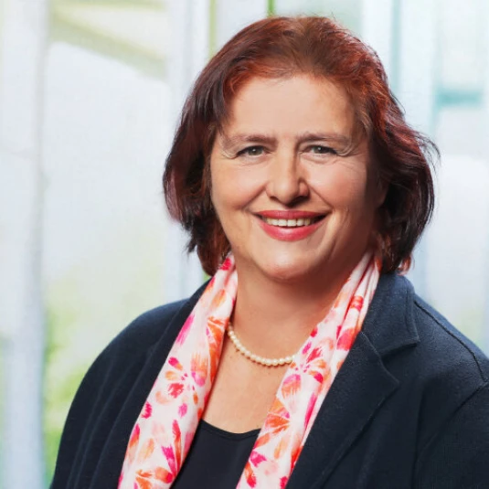 Porträt einer Frau mit schulterlangem braun-rotem Haar. Sie trägt einen dunklen Blazer, eine Halskette und einen bunt gemusterten Schal. Der Hintergrund ist unscharf.