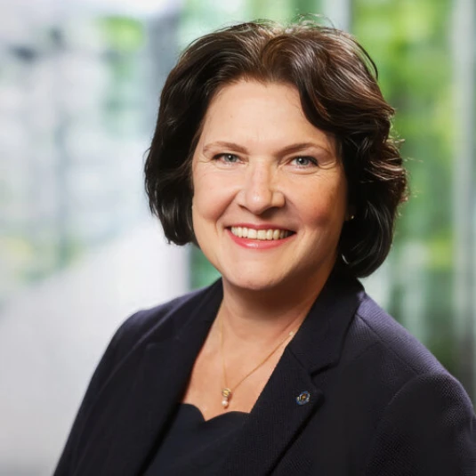 Eine Frau mit schulterlangem, dunklem Haar lächelt in die Kamera. Sie trägt ein dunkelblaues Oberteil und eine feine Halskette. Der Hintergrund ist unscharf und zeigt eine grünliche Umgebung.