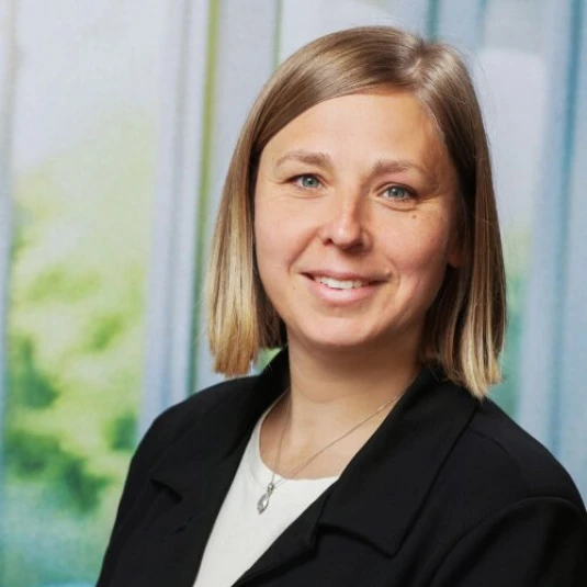 Porträt einer lächelnden Frau mit schulterlangem, glattem Haar. Sie trägt einen schwarzen Blazer und eine Kette mit Anhänger. Der Hintergrund ist unscharf und zeigt helle Farben.
