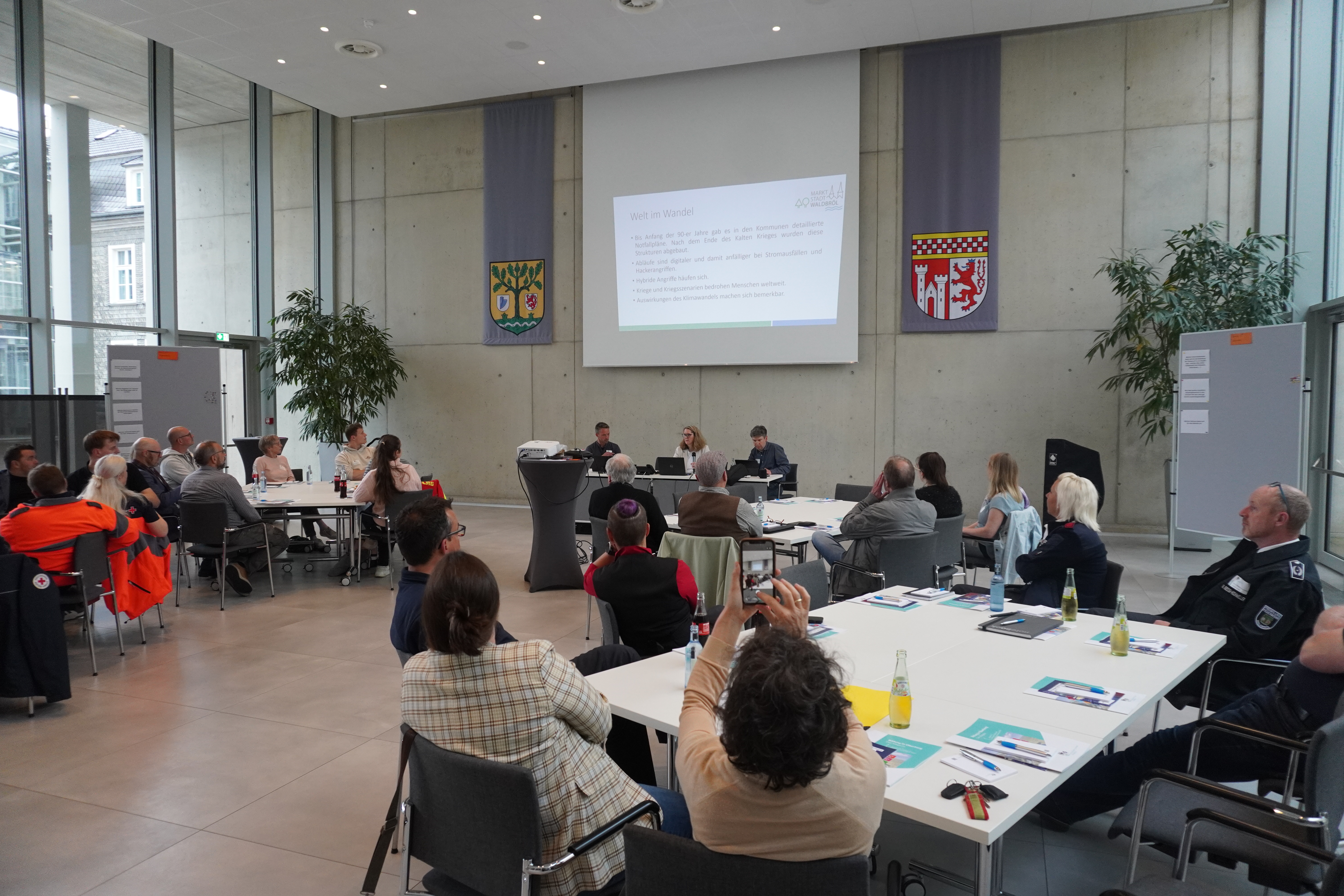 Ein Konferenzraum mit mehreren Personen, die an Tischen sitzen und einer Präsentation folgen. Im Hintergrund ein Projektorbildschirm und zwei Banner an der Wand.